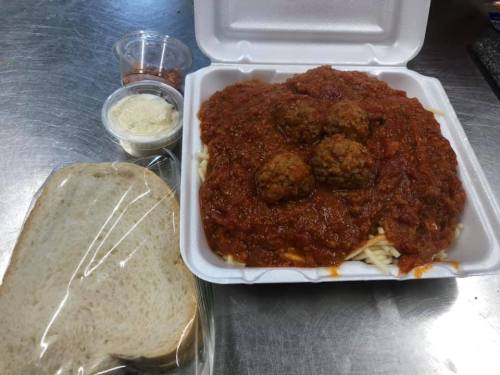 A takeout container with spaghetti and meatballs in marinara sauce, served with a side of bread and condiments.