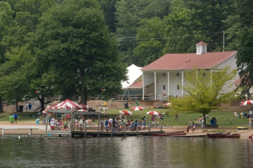 A lakeside scene with a dock, people enjoying the beach, and a white building with a red roof surrounded by trees.