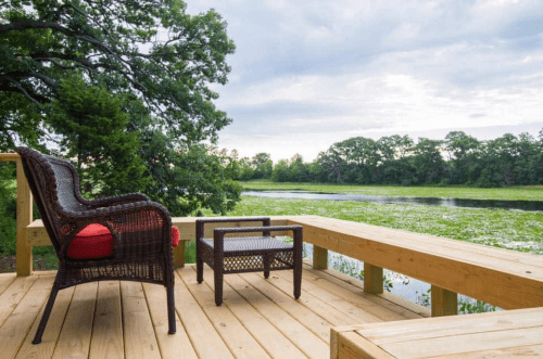 A serene deck overlooking a calm river, featuring wicker chairs and lush greenery in the background.