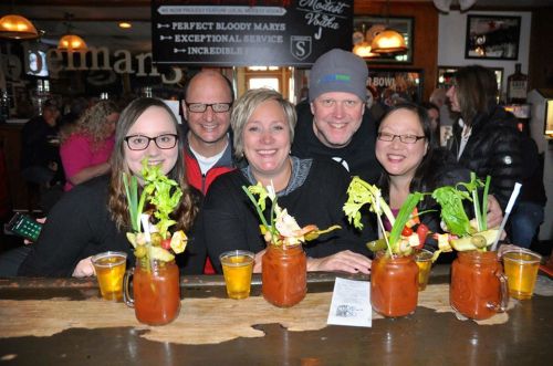 A group of five friends smiles at a bar, each with colorful drinks and garnishes in front of them.