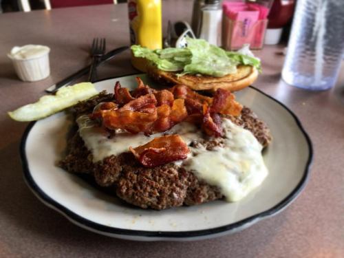 A plate featuring a large burger topped with melted cheese, crispy bacon, lettuce, and a pickle on the side.