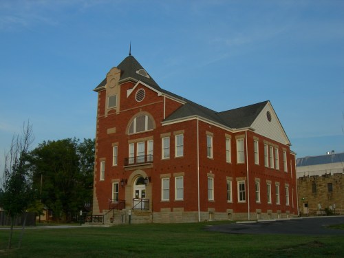 Historic red brick building with a peaked roof, large windows, and a grassy area in front under a clear blue sky.