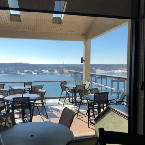A scenic view from a balcony overlooking a lake and bridge, with empty tables and chairs in the foreground.