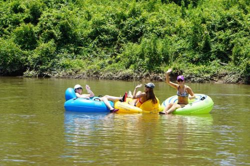 Three people on colorful inner tubes wave while floating on a calm river surrounded by lush greenery.