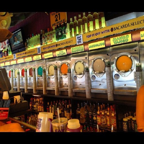 A colorful array of frozen drink machines lined up at a bar, with various flavors displayed above.