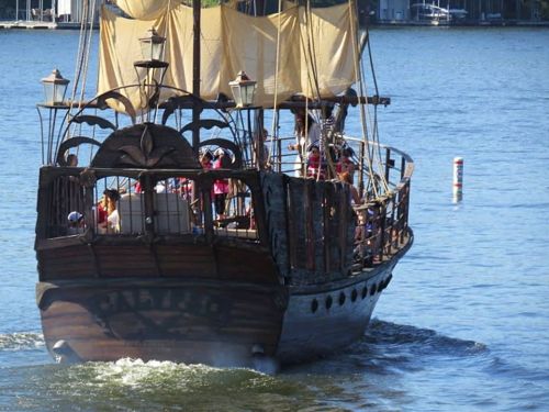 A wooden pirate ship with sails, cruising on a calm lake, filled with passengers enjoying the ride.