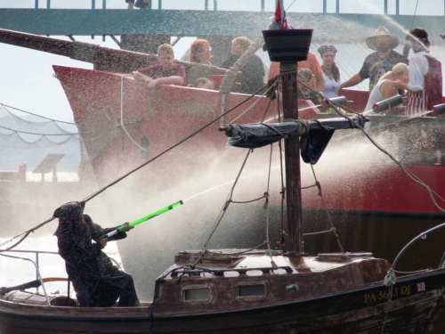 A person in a black outfit sprays water from a boat at a crowd on a larger ship during a lively water battle.