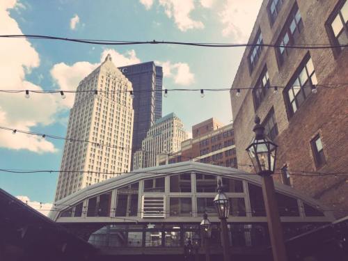 View of city buildings under a blue sky with clouds, featuring string lights and a modern structure in the foreground.