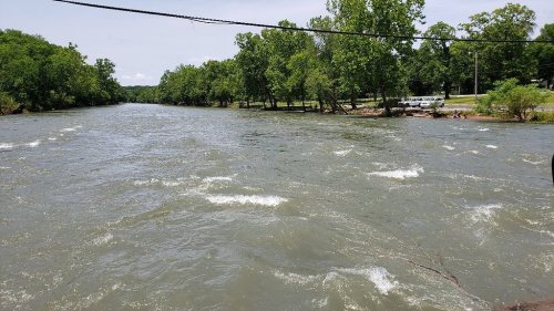 A flowing river surrounded by lush green trees and a grassy bank, with a few people visible in the distance.