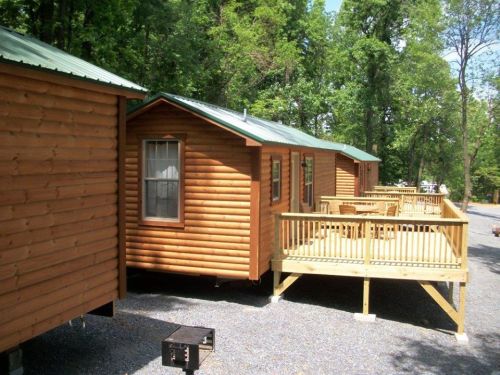 Two wooden cabins with green roofs, surrounded by trees, featuring wooden decks and gravel pathways.