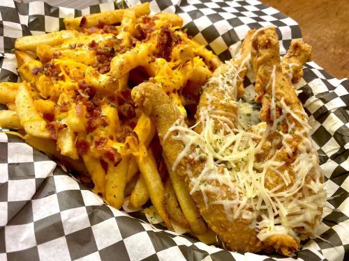 A plate of crispy fried chicken with cheesy fries topped with bacon, served in a checkered paper basket.