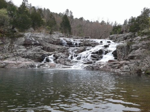 A serene waterfall cascading over rocky terrain into a calm pool, surrounded by trees and a tranquil landscape.