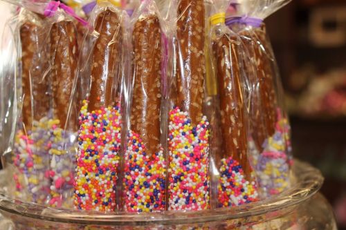 A close-up of chocolate-covered pretzel rods decorated with colorful sprinkles, packaged in clear bags.