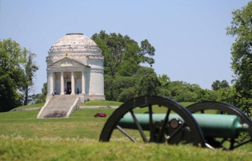 A historic monument with a dome, surrounded by trees, and a cannon in the foreground on a sunny day.