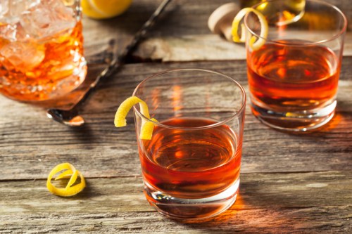 Two glasses of amber-colored cocktails with lemon twists, set on a rustic wooden table. A cocktail shaker is in the background.