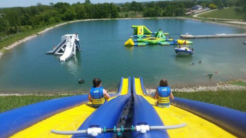 Two children sit on a water slide overlooking a lake with inflatable attractions and a dock in the background.