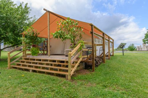 A tent with an orange canopy on a wooden deck, surrounded by greenery and potted plants, under a cloudy sky.
