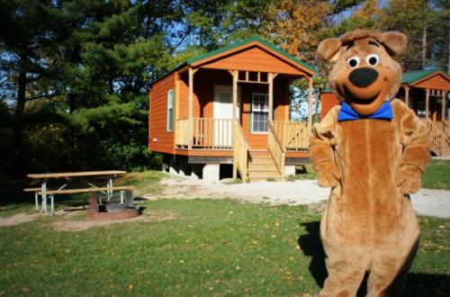 A bear mascot stands in front of cozy cabins with a picnic table and fire pit in a sunny outdoor setting.