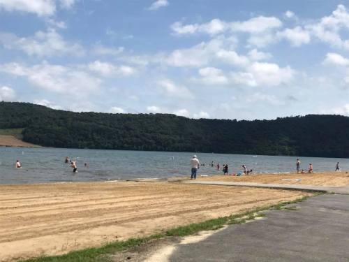 A sandy beach by a lake with people swimming and playing in the water, surrounded by green hills under a cloudy sky.