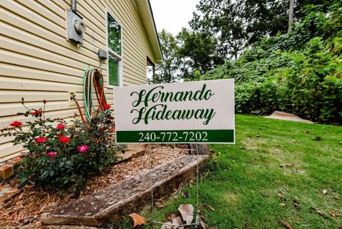 Sign for "Hernando Hideaway" with a phone number, placed in a garden area with flowers and greenery in the background.