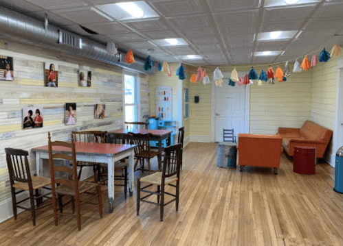 Brightly lit room with wooden floors, colorful wall decor, a dining table, chairs, and a cozy seating area.