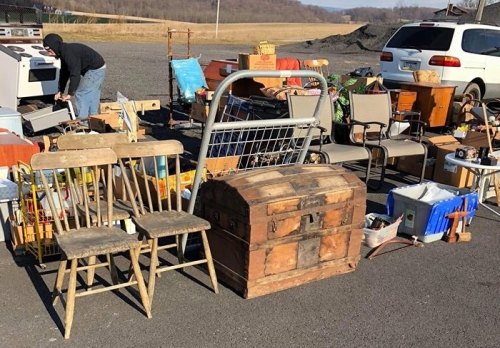 A variety of secondhand items for sale, including chairs, a wooden chest, and household goods, at an outdoor market.