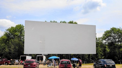 A blank outdoor movie screen surrounded by cars and people enjoying a sunny day. Trees in the background.
