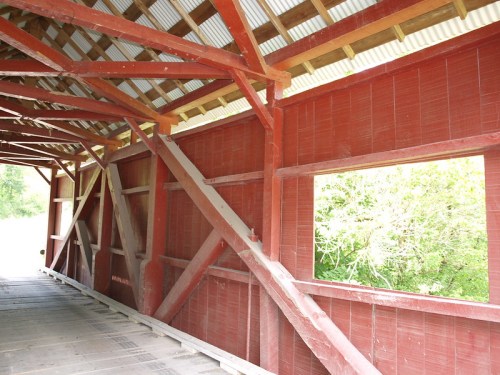 Interior view of a red covered bridge, showcasing wooden beams and an open window with greenery outside.