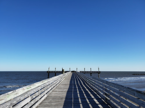 A wooden pier extends into the calm ocean under a clear blue sky, with gentle waves lapping at the shore.