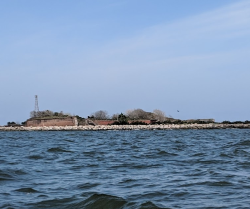 A distant view of a rocky shoreline with an old fort and a tower against a clear blue sky. Waves in the foreground.