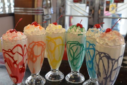 Six colorful milkshakes topped with whipped cream and cherries, arranged in tall glasses on a counter.