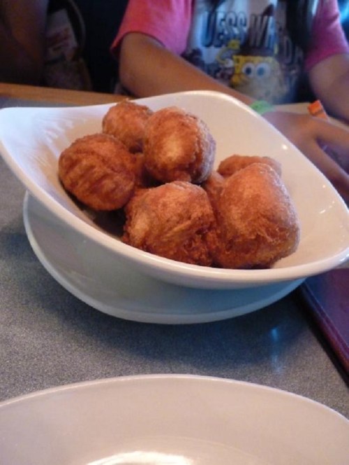 A bowl of golden-brown fried dough balls on a table, with hands and a colorful shirt in the background.