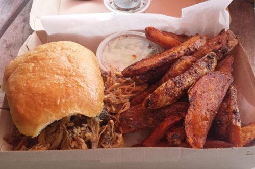 A box containing a pulled pork sandwich and crispy sweet potato fries, with a small container of coleslaw.