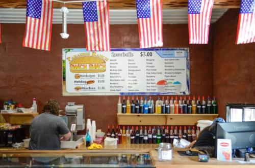 A food stand with a menu featuring hamburgers, hotdogs, nachos, and snowballs, decorated with American flags.