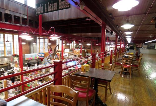 A view of a bustling food market from an upper level, featuring wooden tables and colorful vendor stalls below.