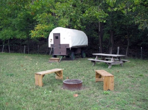 A covered wagon parked in a grassy area, surrounded by wooden benches and a picnic table near a fire pit.