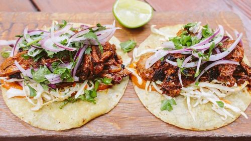 Two tacos on a wooden board, topped with shredded meat, cabbage, onions, and cilantro, with a lime wedge on the side.