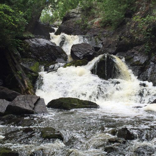 A cascading waterfall flows over rocky terrain, surrounded by lush green trees and foliage.