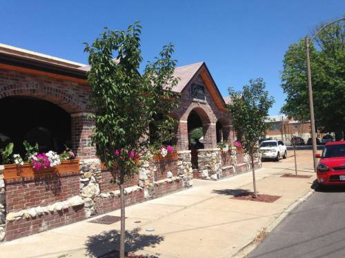 A brick building with arched windows, surrounded by trees and flowers, on a sunny day. Cars parked nearby.