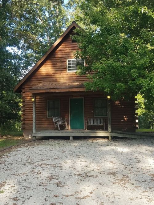 A rustic log cabin with a green door, surrounded by trees, featuring two rocking chairs on the porch.