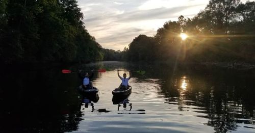 Two people kayaking on a calm river at sunset, surrounded by trees and reflecting water.