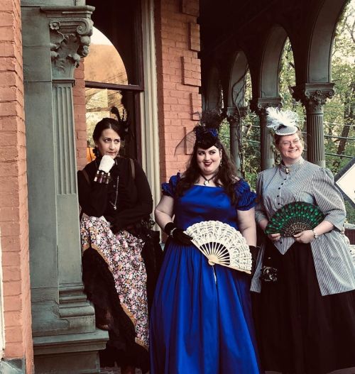 Three women in vintage dresses and accessories pose on a porch, showcasing elegant fans and historical fashion.