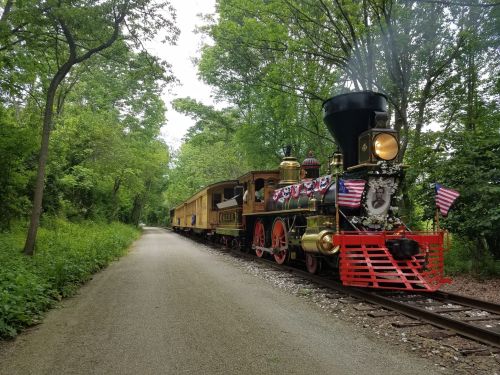 A vintage steam train decorated with flags, parked on a tree-lined track beside a gravel path.