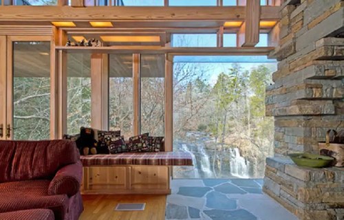 Cozy living room with a stone wall, large windows, and a view of a waterfall surrounded by trees.