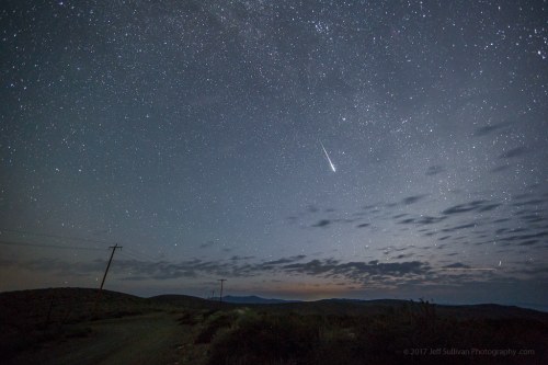 A starry night sky with a shooting star over a remote landscape and distant mountains.