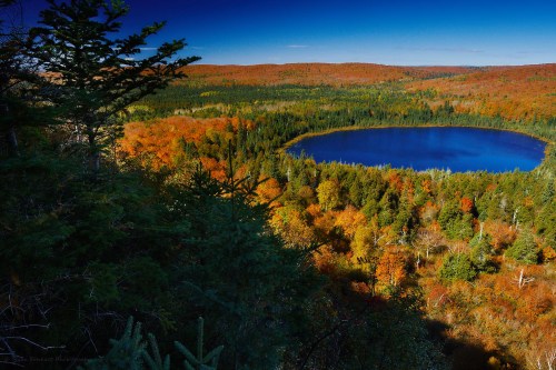 A vibrant autumn landscape featuring a blue lake surrounded by colorful fall foliage and evergreen trees.