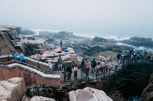 A crowd of people walking along a rocky coastal path with fog and ocean waves in the background.