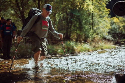 A hiker crosses a stream using trekking poles, surrounded by lush greenery and fellow hikers in the background.