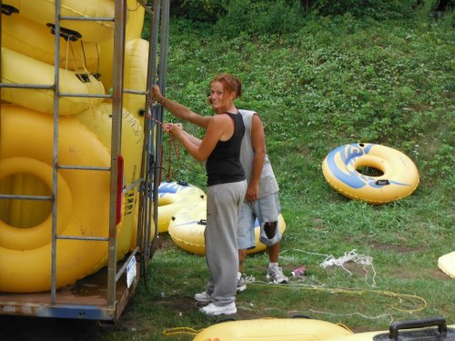Two people unloading yellow inflatable tubes from a trailer, with more tubes scattered on the grass in the background.