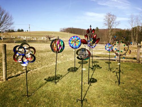 Colorful wind sculptures on metal stands in a grassy field, with a cemetery and hills in the background.